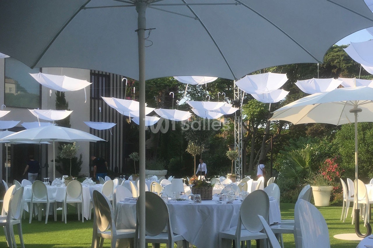Tables dressées en extérieur sous des parasols blancs, avec des parapluies suspendus en décoration.