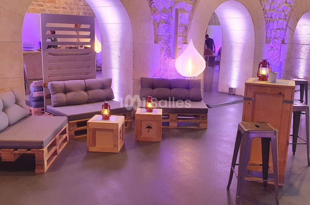 Espace lounge avec canapés en palettes, tables basses en bois et éclairage tamisé dans une salle voûtée en pierre.