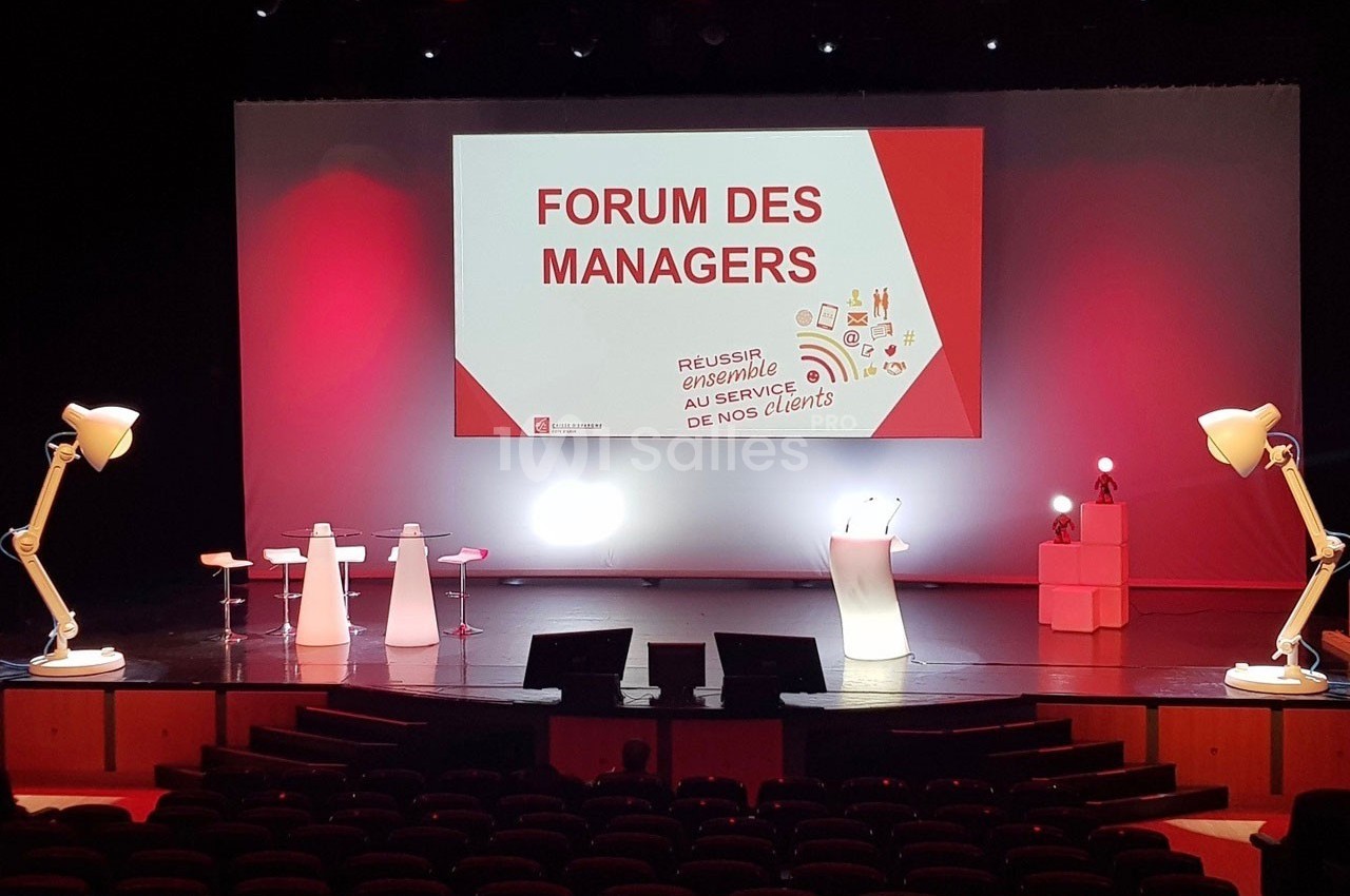 Scène de conférence avec écran affichant ’Forum des Managers’, pupitre, tabourets et éclairage rouge et blanc.