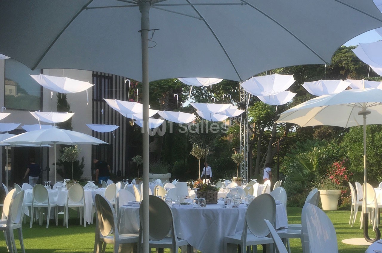 Tables dressées en extérieur sous des parasols blancs, avec des parapluies suspendus en décoration.