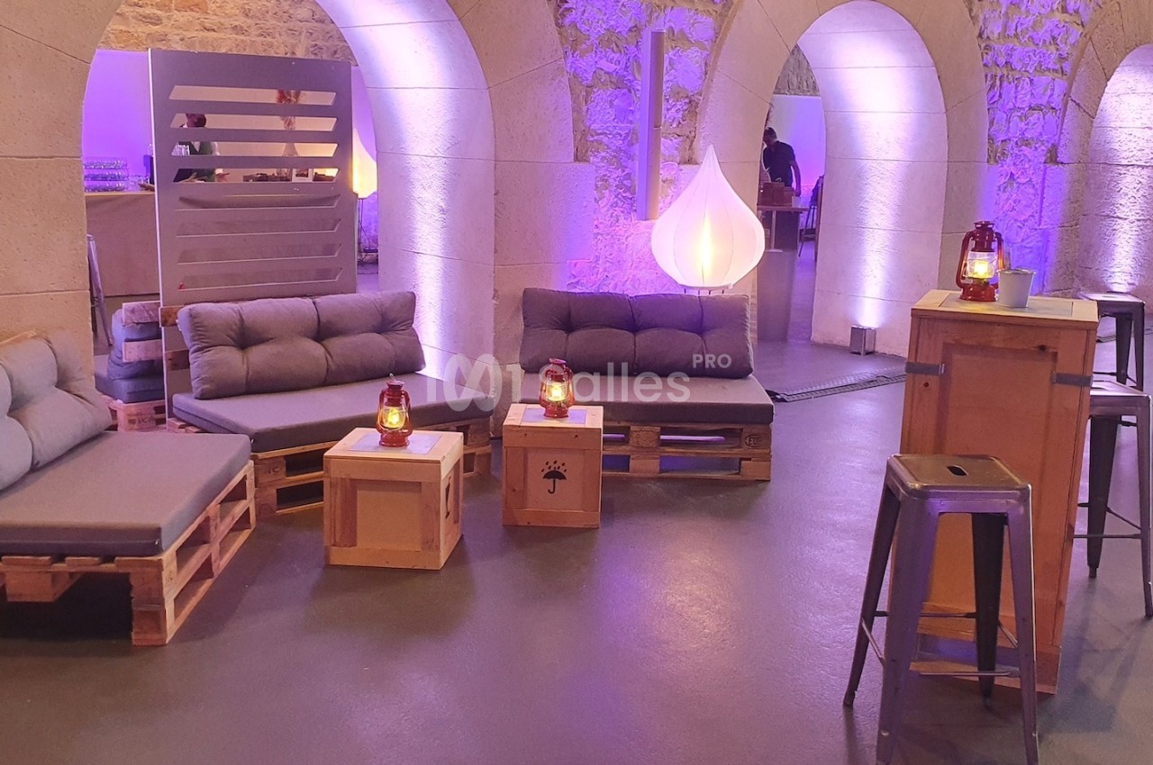 Espace lounge avec canapés en palettes, tables basses en bois et éclairage tamisé dans une salle voûtée en pierre.