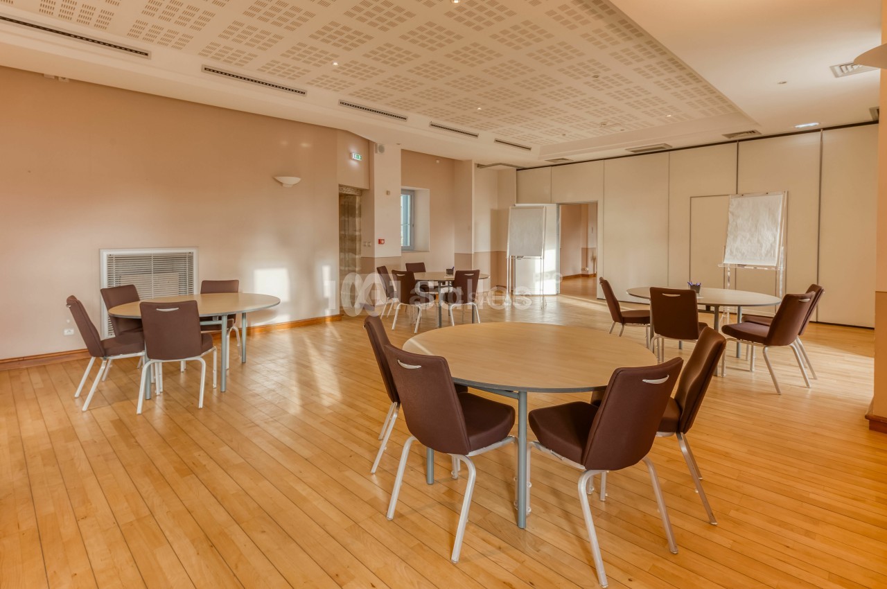 Salle lumineuse avec parquet, tables rondes et chaises marron, équipée de panneaux et tableaux blancs.