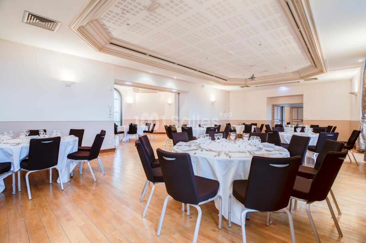 Salle de réception lumineuse avec tables rondes dressées, chaises noires et parquet en bois clair.