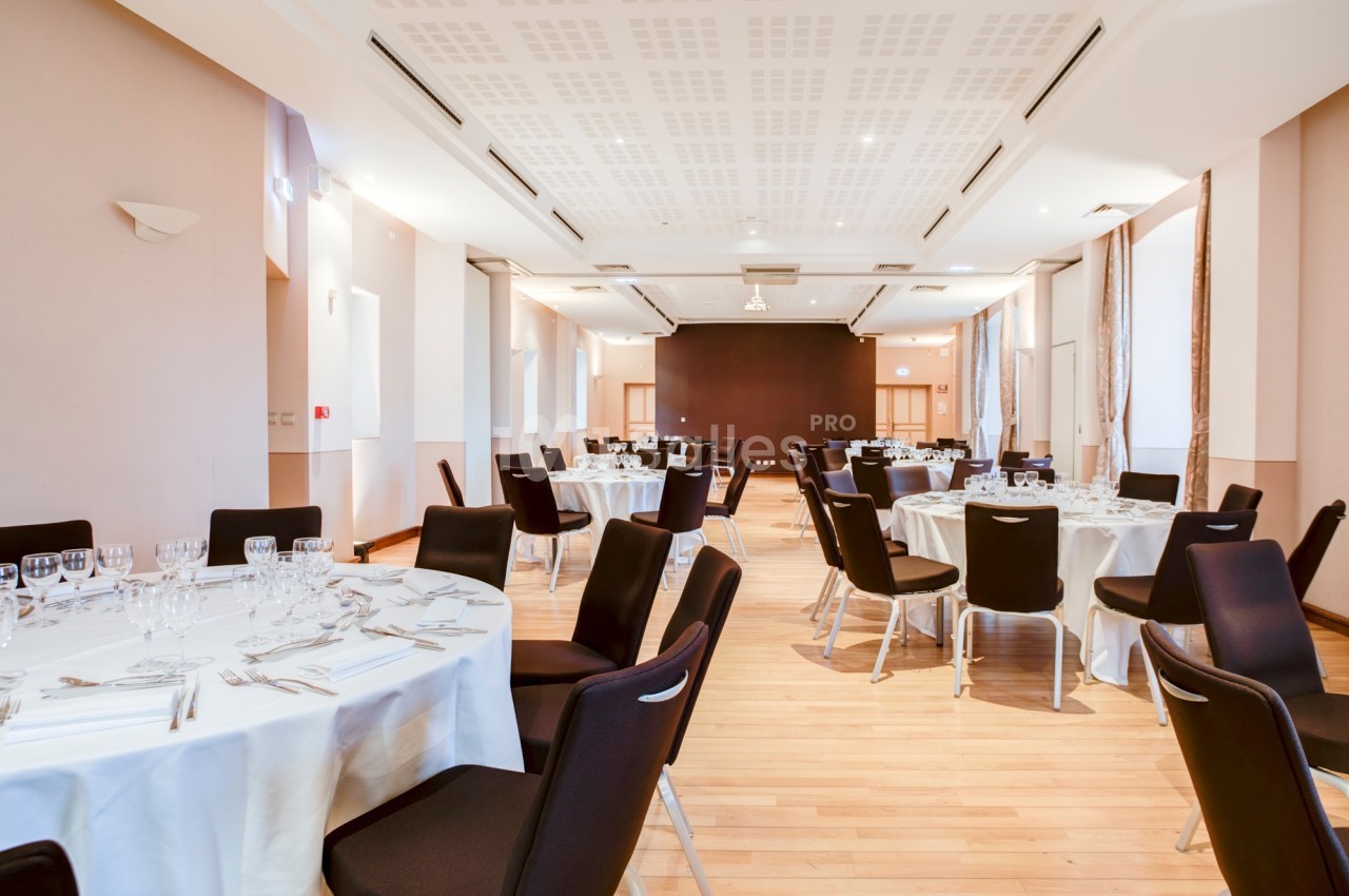 Salle de réception lumineuse avec tables rondes dressées, chaises noires et parquet clair.