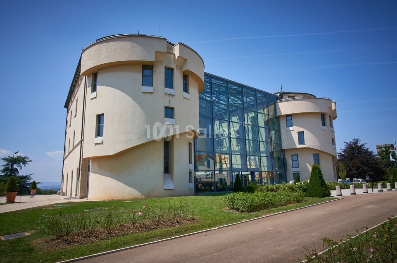 Bâtiment moderne avec façade en verre et murs arrondis, entouré de pelouse et d'arbustes sous un ciel bleu.