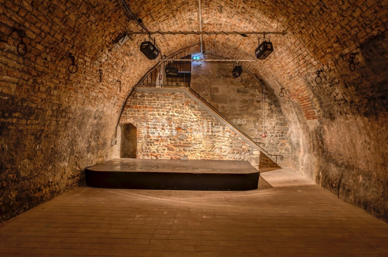 Salle voûtée en briques avec une petite scène surélevée, éclairée par des projecteurs suspendus.