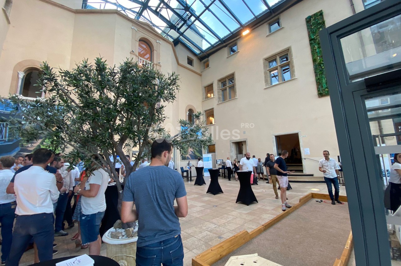 Personnes rassemblées dans une cour intérieure lumineuse avec un toit en verre, un terrain de pétanque et des tables hautes.
