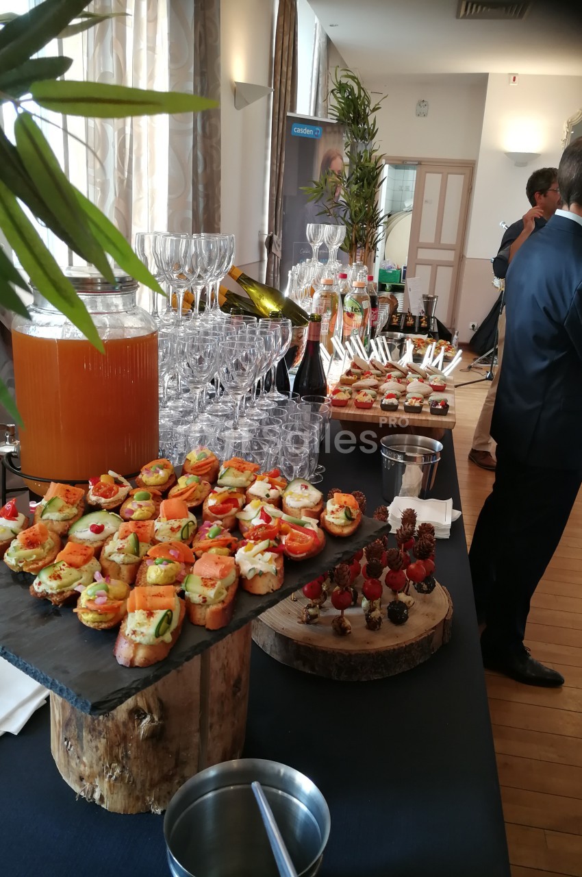 Buffet avec verrines, canapés variés, brochettes de fruits et jus disposés sur une table dans une salle lumineuse.