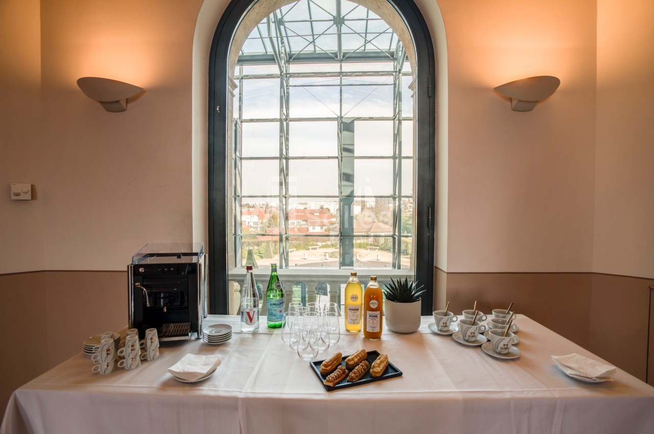 Table dressée avec machine à café, bouteilles, verres, tasses et viennoiseries, devant une grande fenêtre lumineuse.