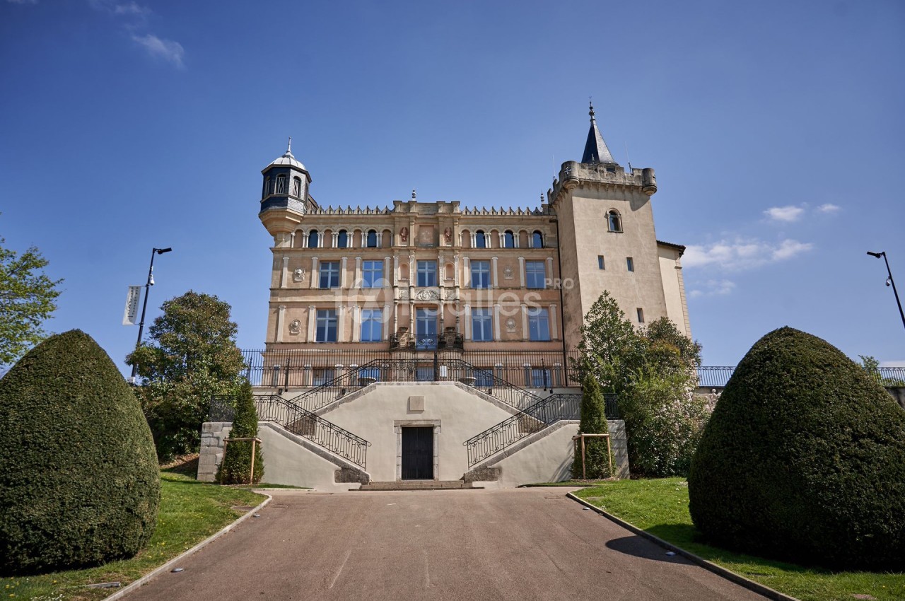Façade d'un château avec escaliers symétriques, tourelles et jardin arboré sous un ciel bleu.