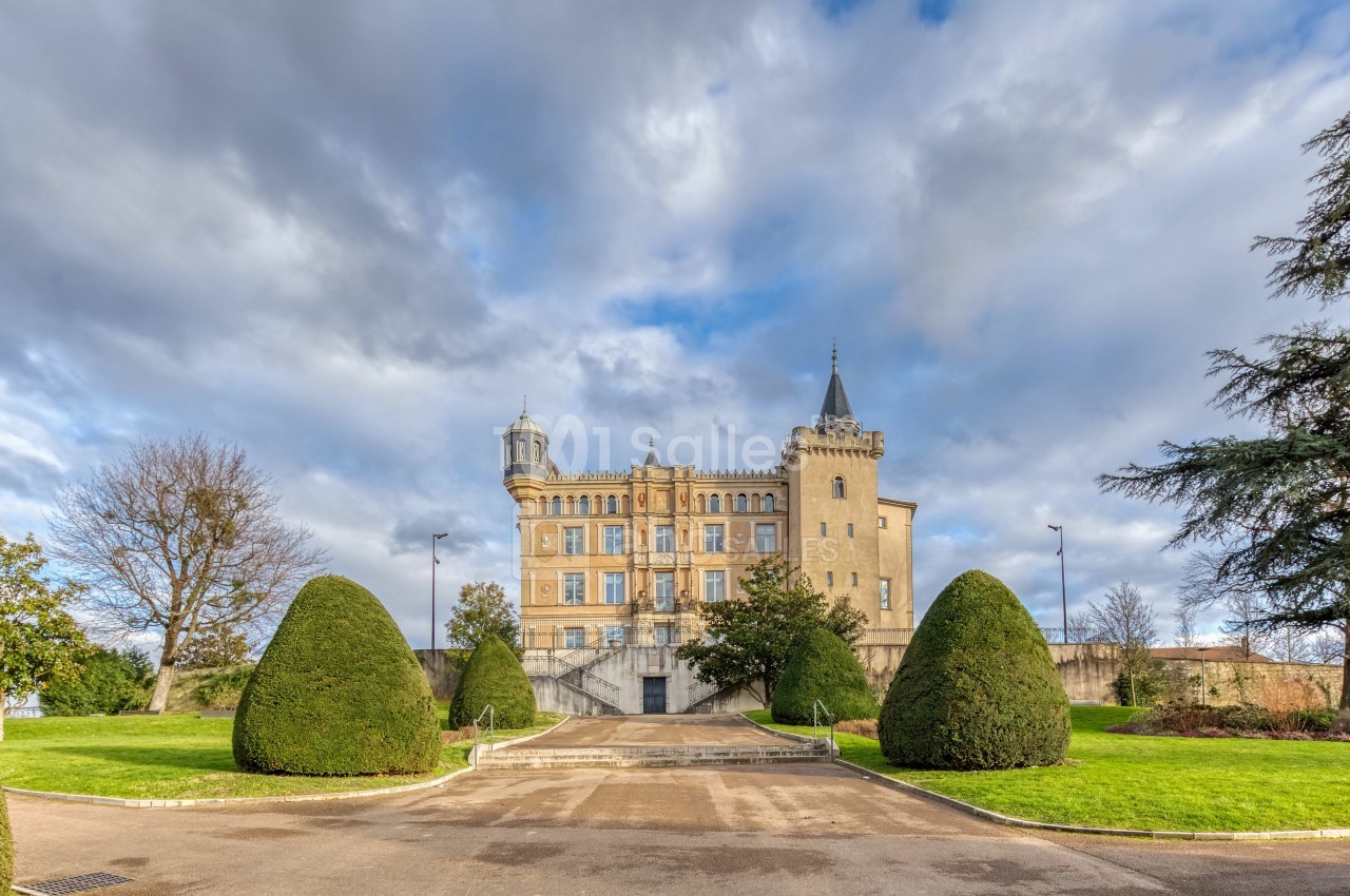 Façade d'un château historique entouré de pelouses bien entretenues et d'arbres taillés, sous un ciel nuageux.