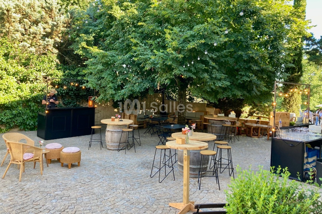 Terrasse extérieure avec tables hautes, chaises en rotin et bar sous des arbres, éclairée par des guirlandes lumineuses.