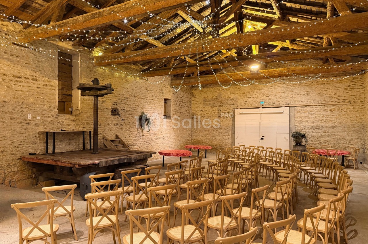 Salle en pierre avec charpente apparente, éclairée par des guirlandes lumineuses, aménagée avec des chaises en bois.