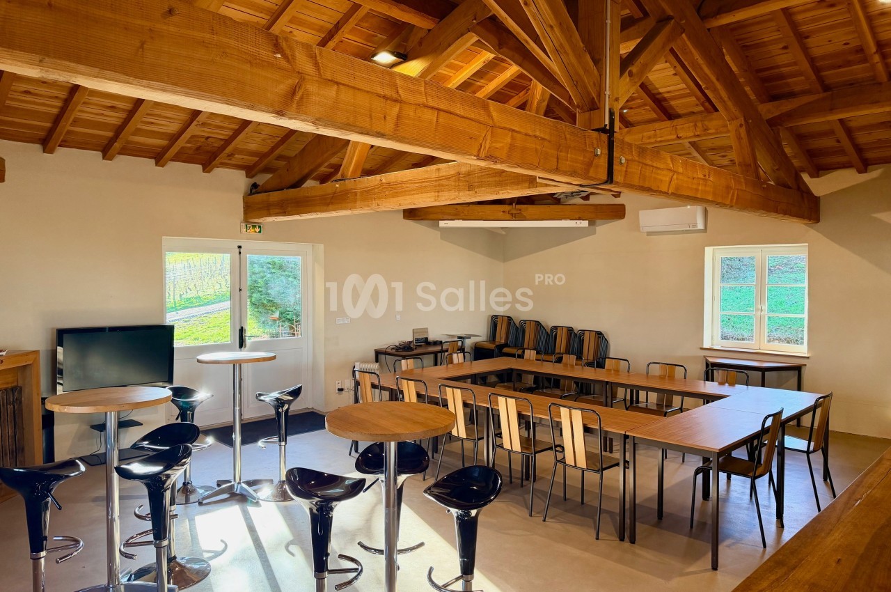 Salle lumineuse avec charpente en bois, tables, chaises et tabourets, donnant sur un extérieur verdoyant.
