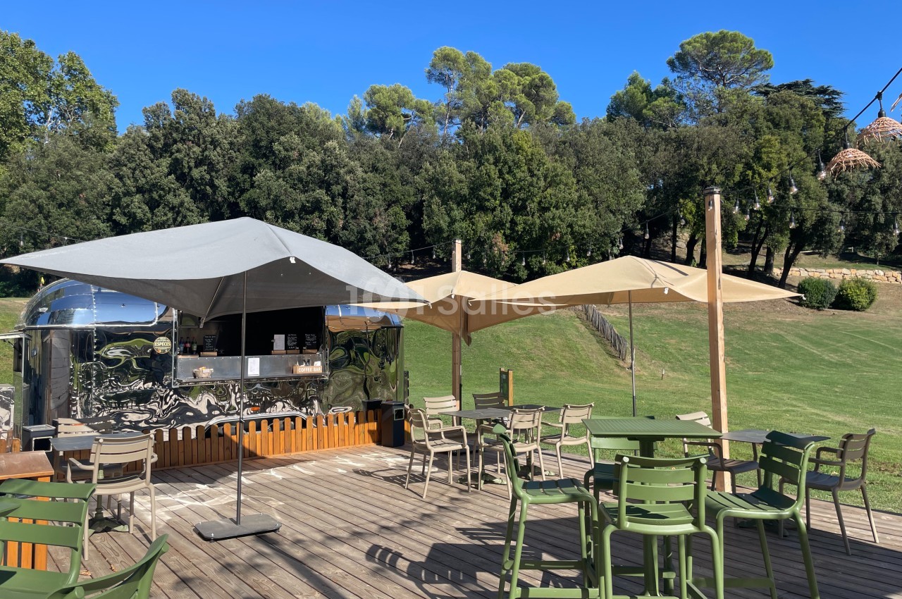 Terrasse en bois avec des tables, des chaises, des parasols et un food truck argenté, entourée de verdure.