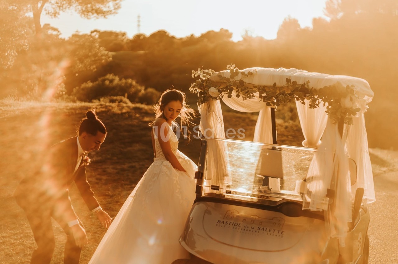 Un couple en tenue de mariage près d'une voiturette de golf décorée, éclairé par une lumière dorée au coucher du soleil.