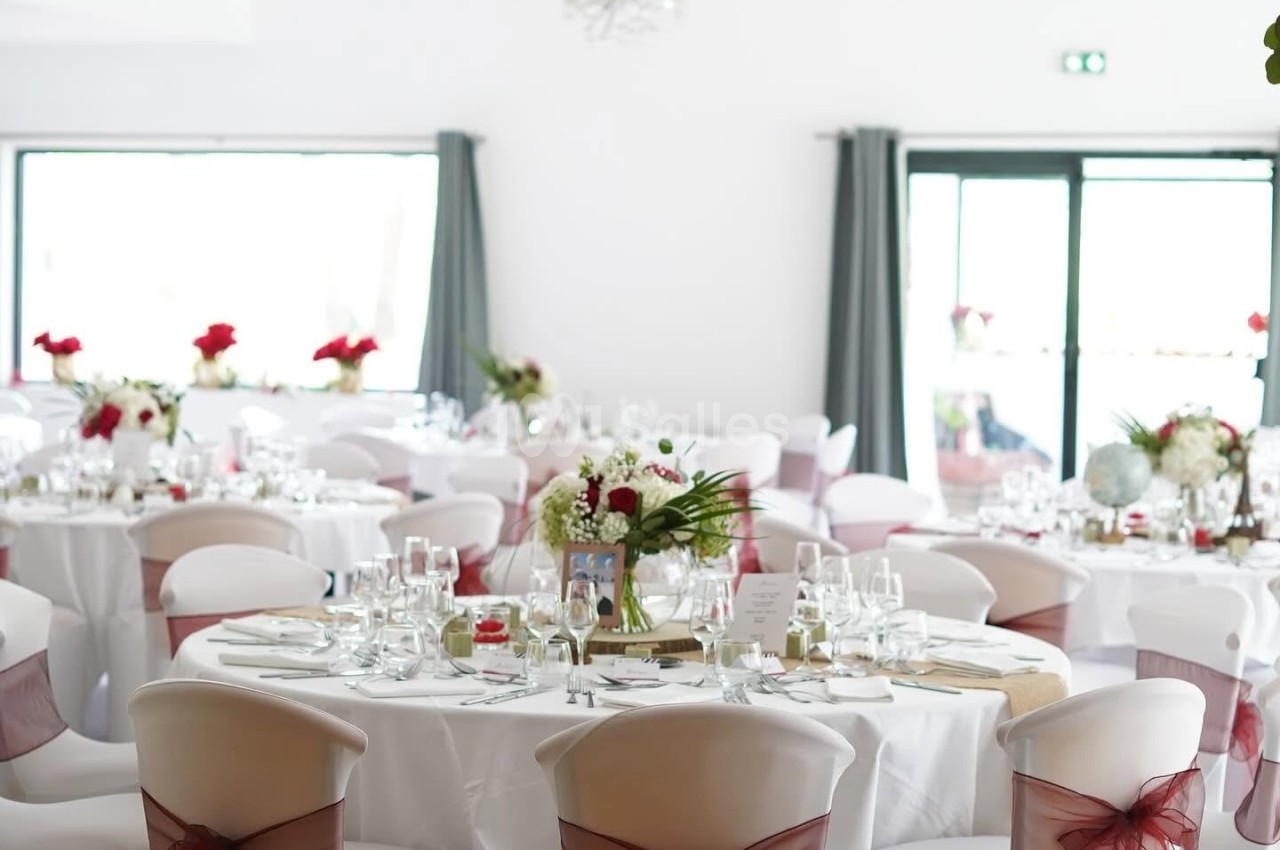 Salle de réception lumineuse avec tables rondes décorées de nappes blanches, rubans rouges et bouquets de fleurs.