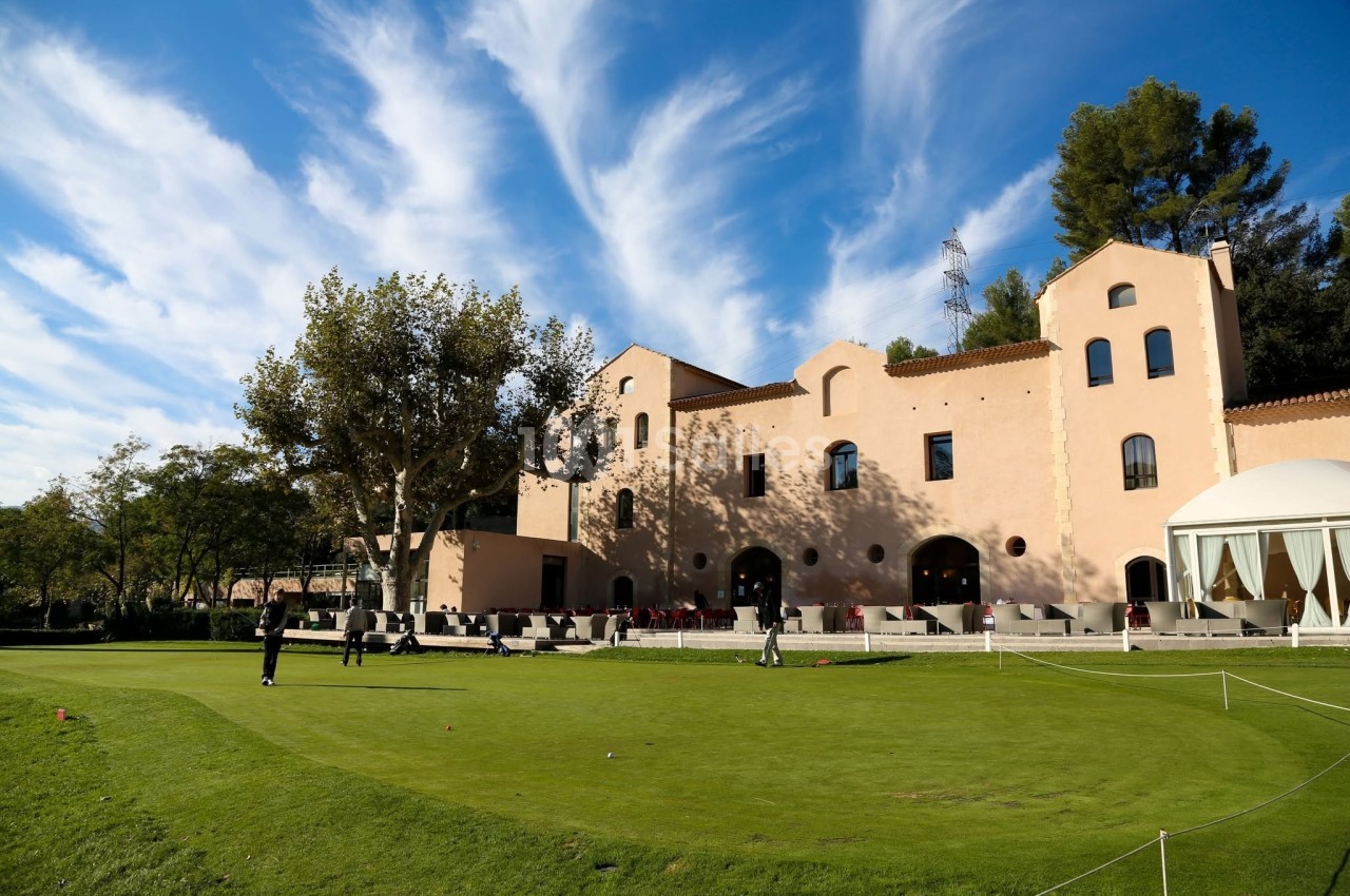 Bâtiment beige entouré d'arbres avec un terrain de golf au premier plan sous un ciel bleu parsemé de nuages.