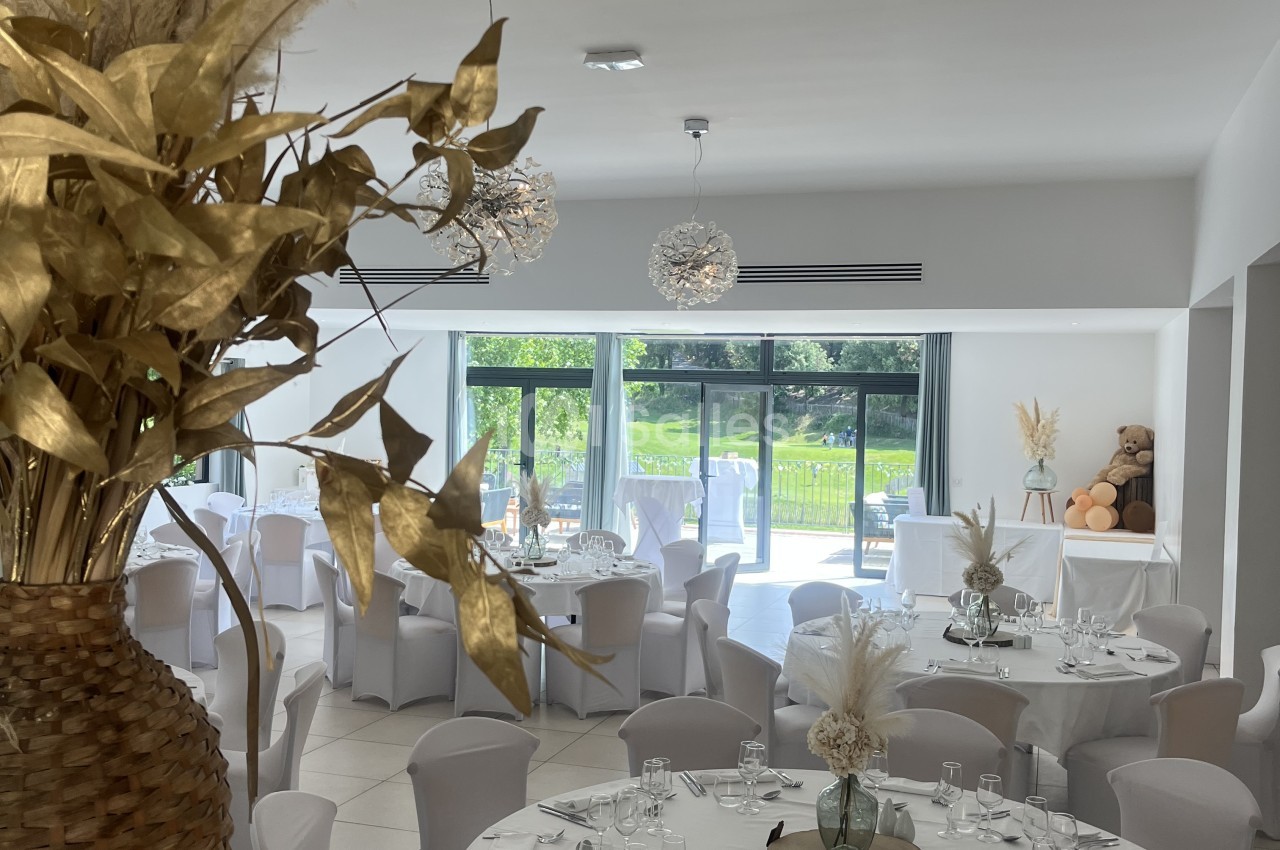 Salle de réception blanche avec tables dressées, chaises grises et baies vitrées ouvrant sur jardin verdoyant.