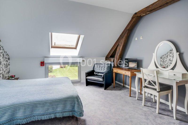 Chambre mansardée avec lit, fauteuil, coiffeuse, bureau, mur bleu, poutre apparente et fenêtre donnant sur un jardin.
