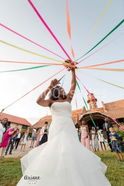 Une mariée en robe blanche tient un bouquet relié à des rubans colorés, entourée d'invités dans un cadre champêtre.