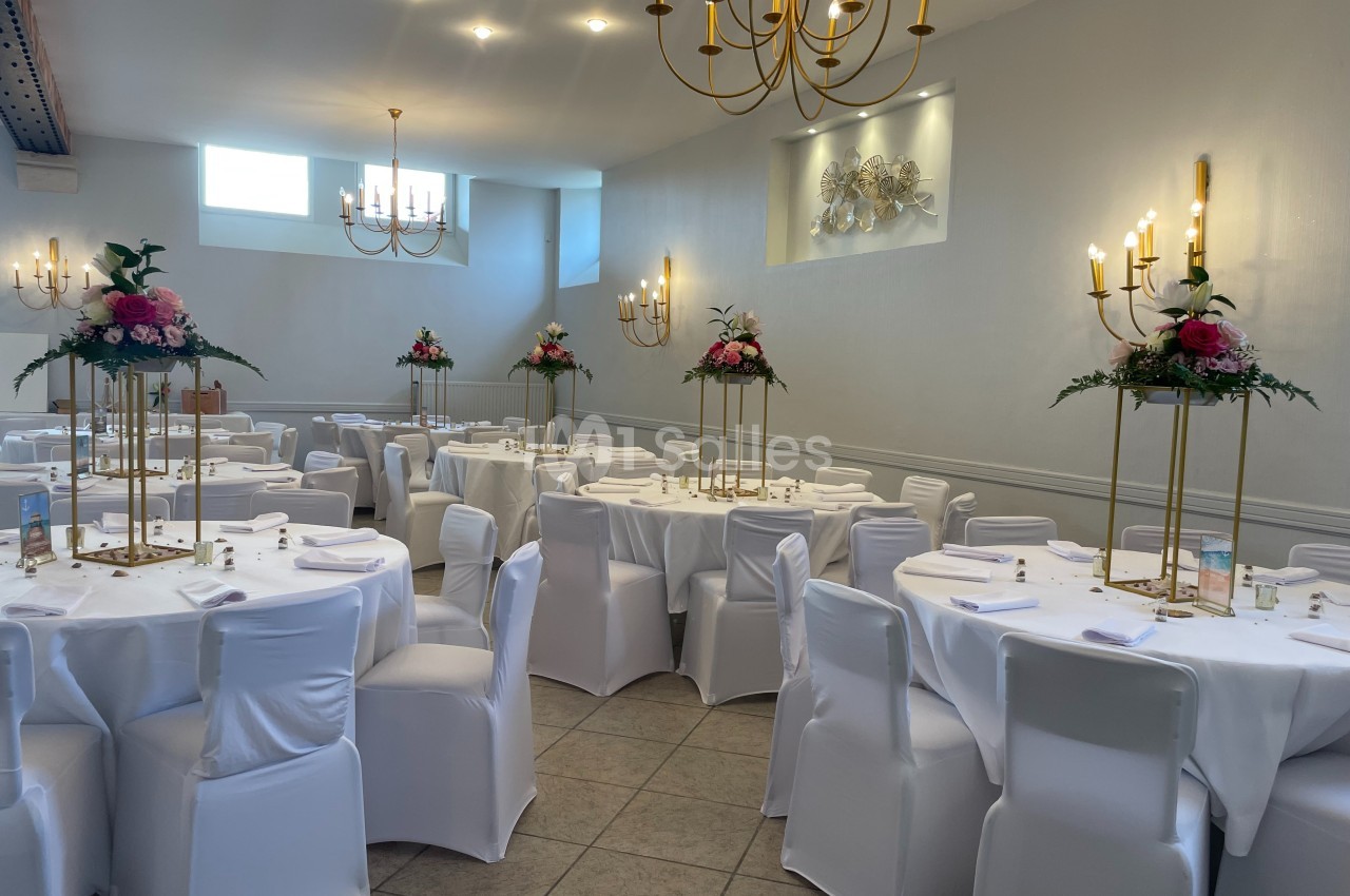 Salle de réception élégante avec tables rondes dressées, nappes blanches et décorations florales sur supports dorés.