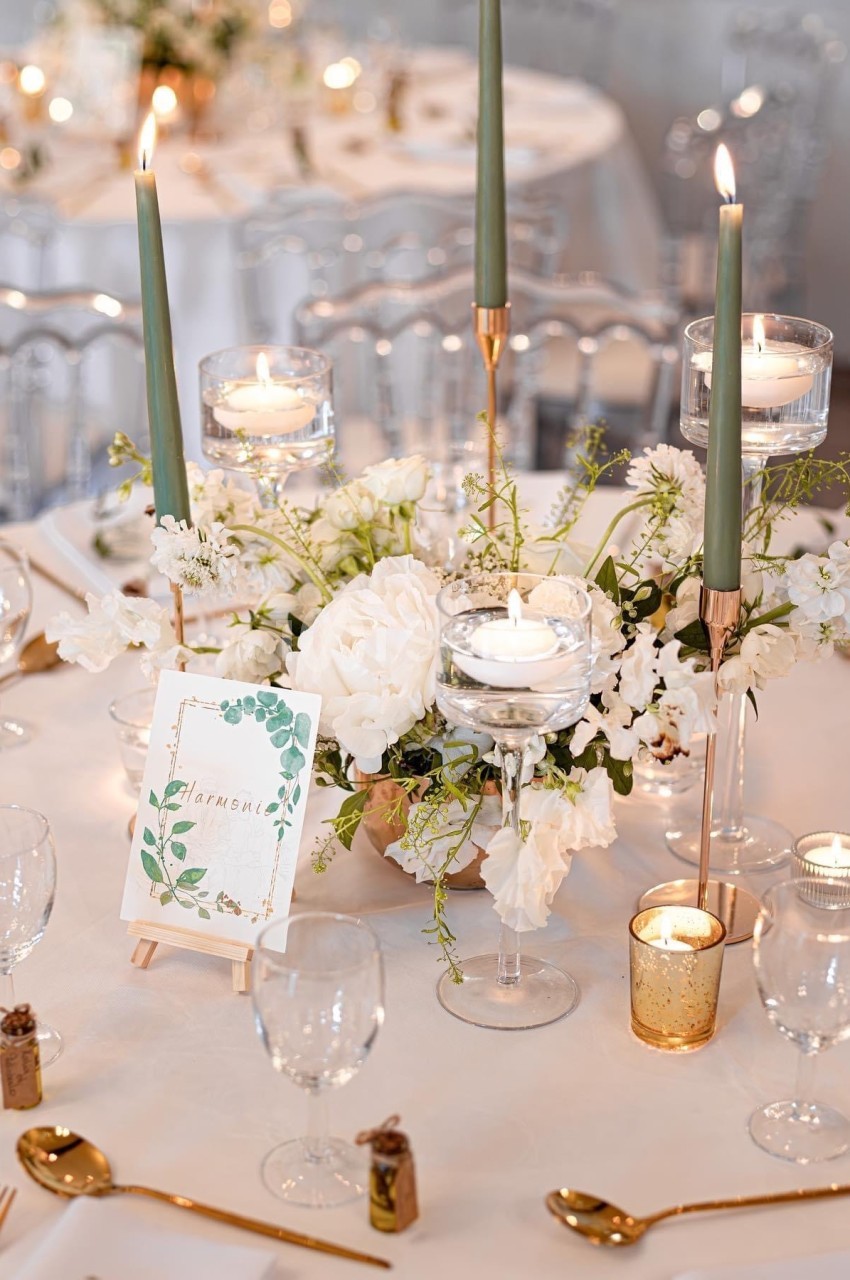 Table élégamment dressée avec bougies, fleurs blanches, verres et carte décorative sur un chevalet.