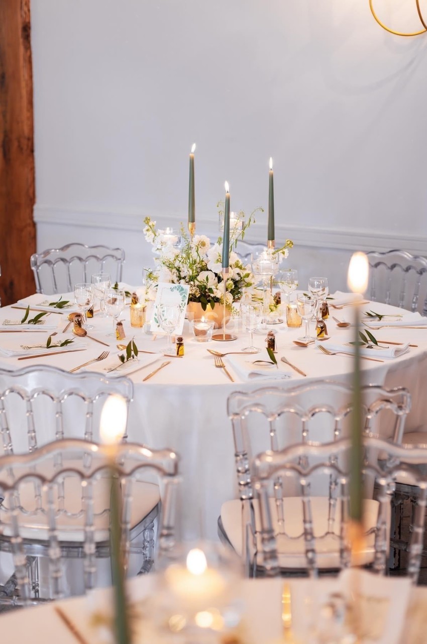 Table ronde décorée pour un événement, avec nappes blanches, bougies allumées et chaises transparentes.