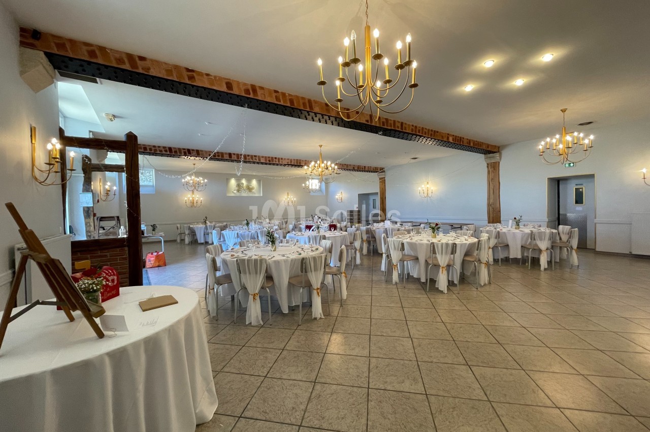 Salle de réception lumineuse avec tables rondes dressées, décorée de lustres et poutres apparentes.