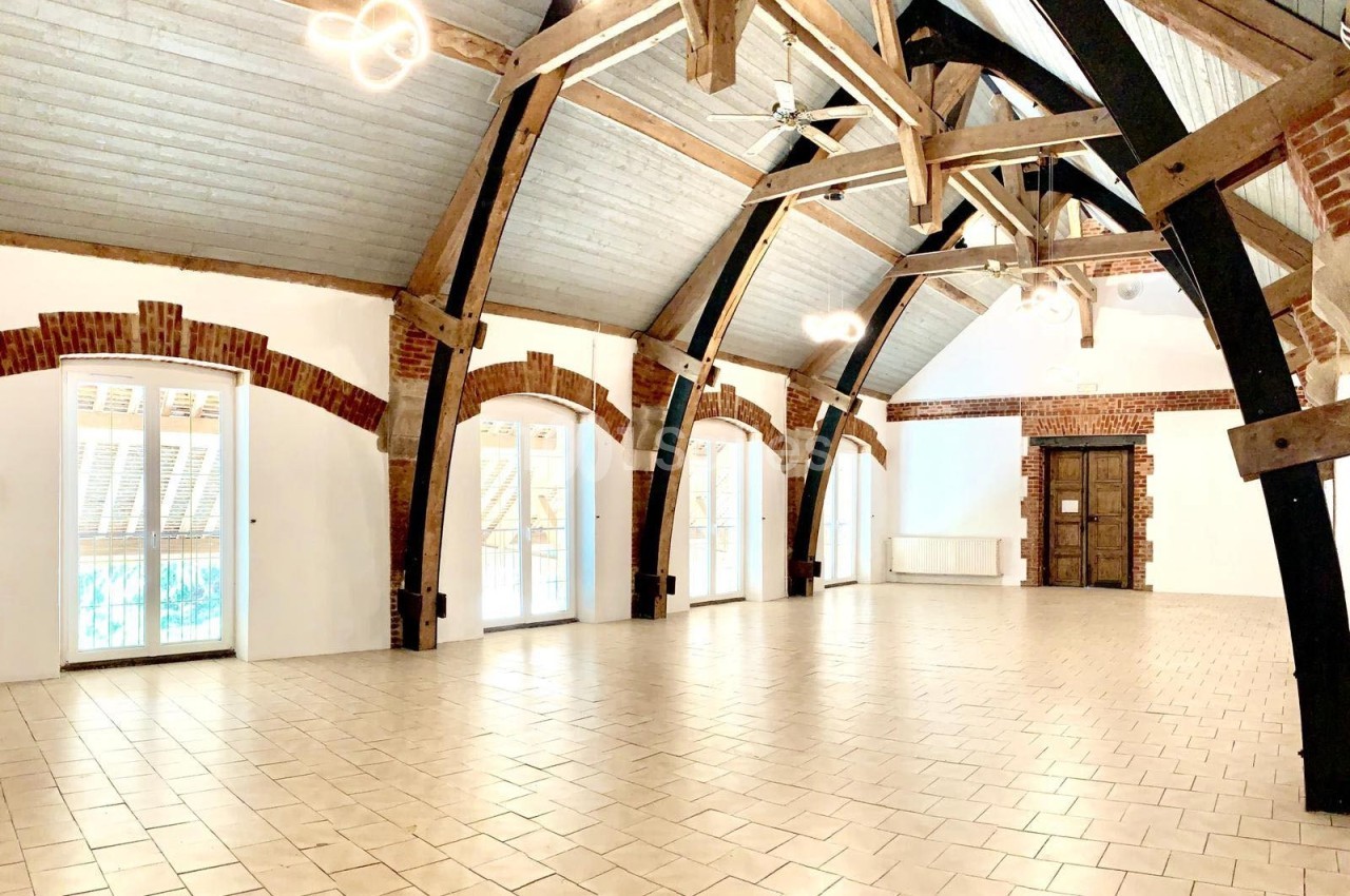 Salle spacieuse avec poutres apparentes, murs en briques et grandes fenêtres laissant entrer la lumière naturelle.