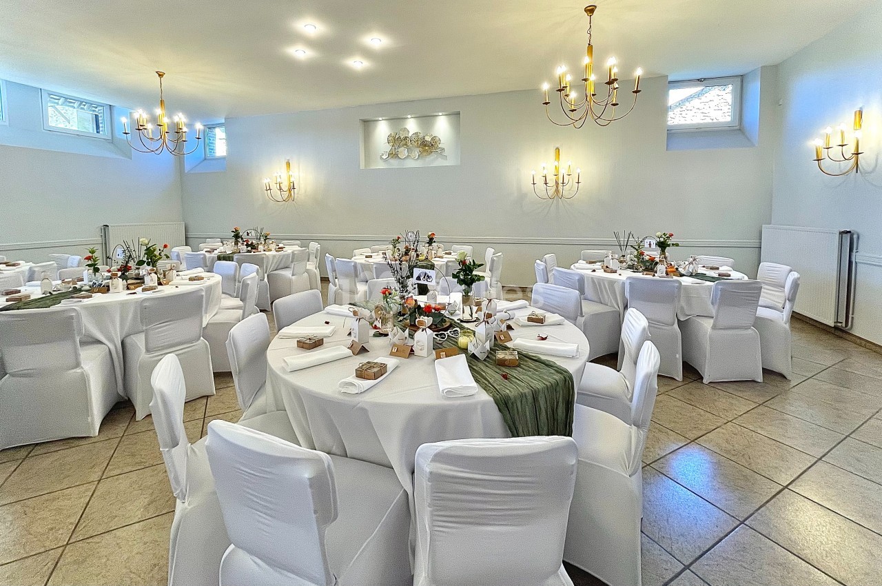 Salle de réception lumineuse avec tables rondes décorées de nappes blanches, centres de table floraux et chandeliers.