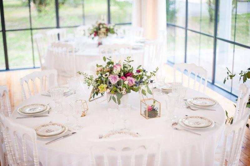 Table ronde décorée avec une nappe blanche, un centre de table floral et de la vaisselle élégante dans une salle lumineuse.