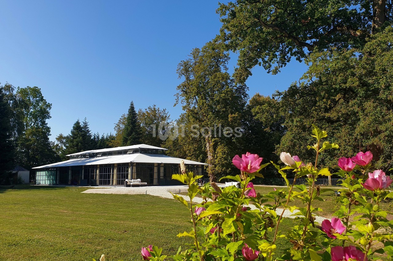Bâtiment moderne avec de grandes baies vitrées entouré d'un jardin verdoyant et de fleurs roses au premier plan.