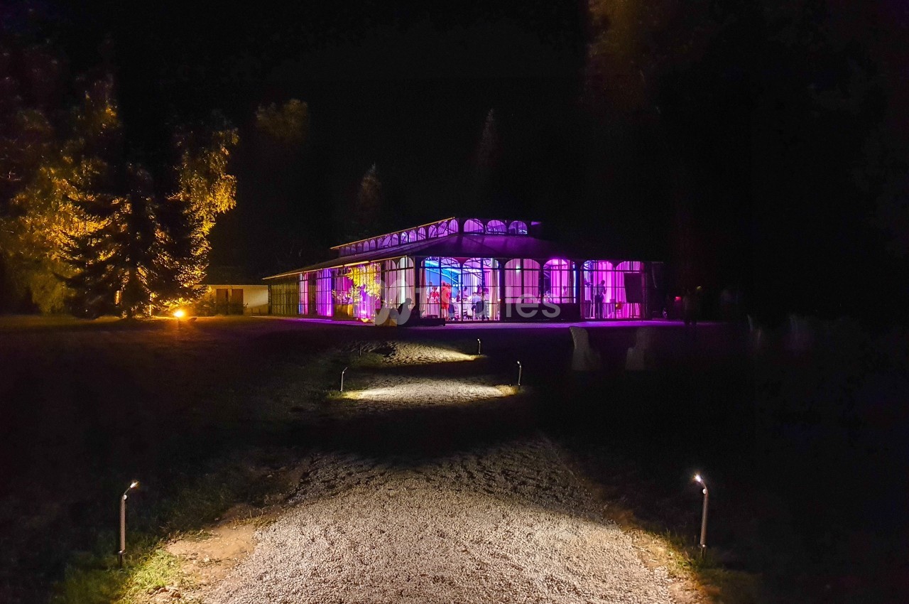 Chemin éclairé menant à un bâtiment avec façade vitrée illuminée de lumières colorées dans un cadre nocturne.