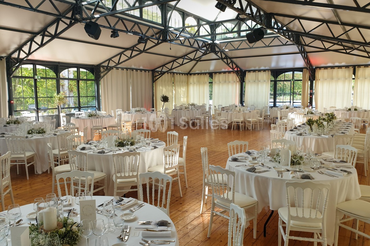 Salle de réception lumineuse avec tables rondes dressées, chaises blanches et grandes baies vitrées donnant sur un espace…
