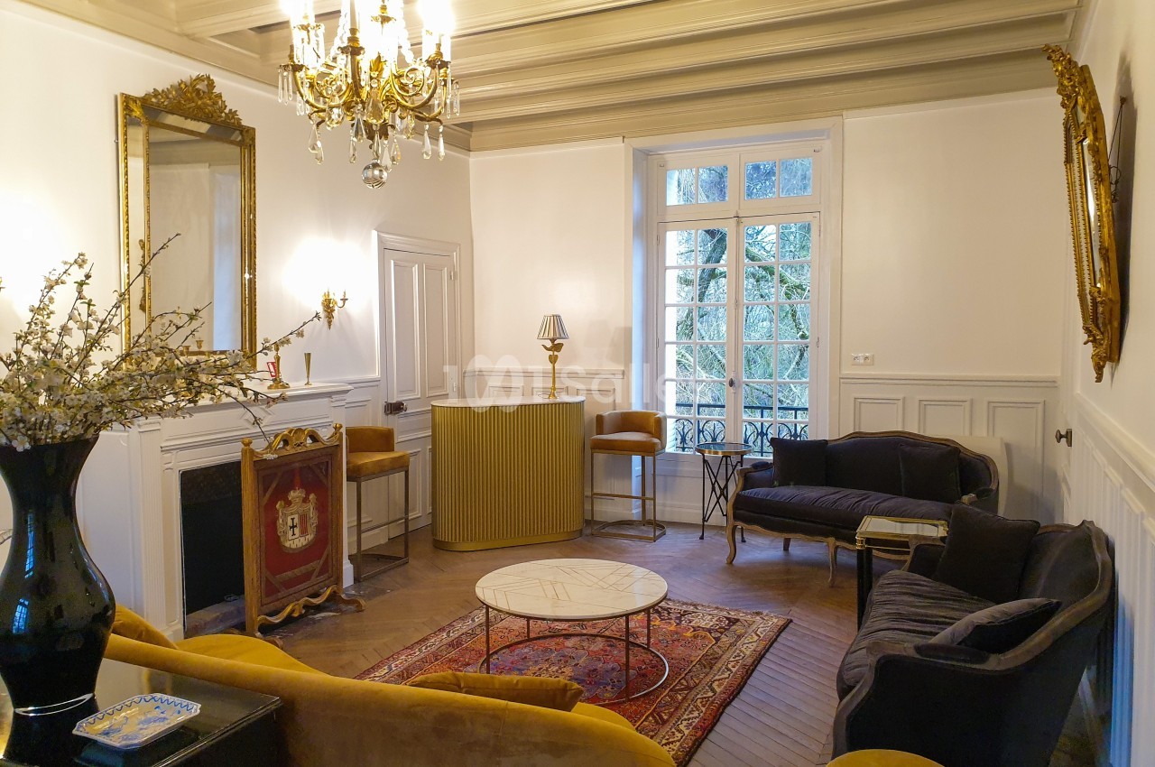 Salon élégant avec mobilier vintage, cheminée, lustre doré, miroirs et grandes fenêtres laissant entrer la lumière naturelle.