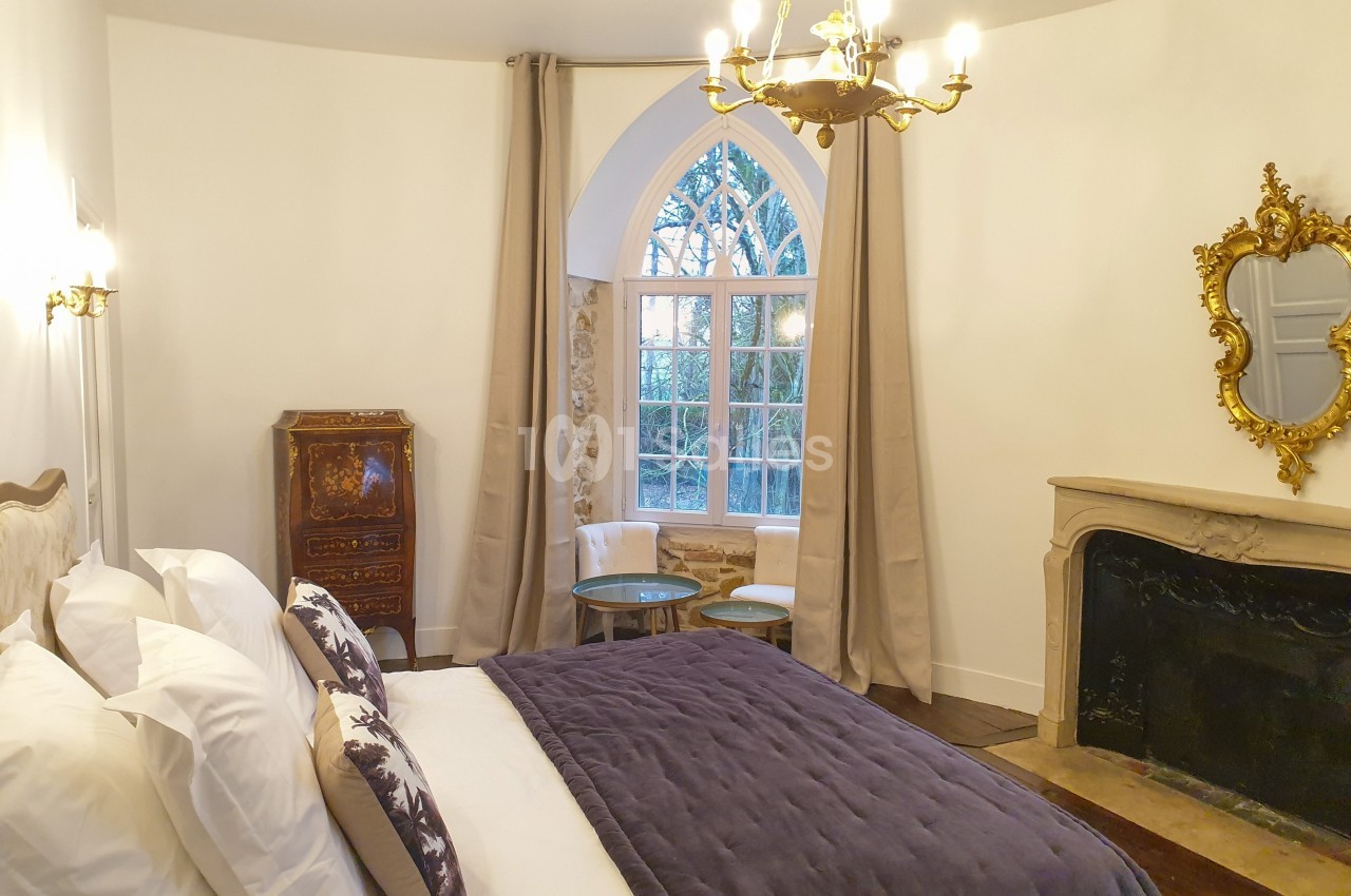Chambre lumineuse avec lit double, cheminée en pierre, miroir doré, meuble ancien et grande fenêtre en arche.
