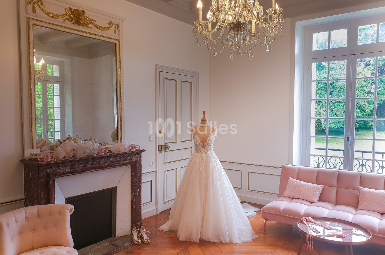 Robe de mariée exposée sur un mannequin dans un salon élégant avec cheminée, lustre et grandes fenêtres donnant sur un…
