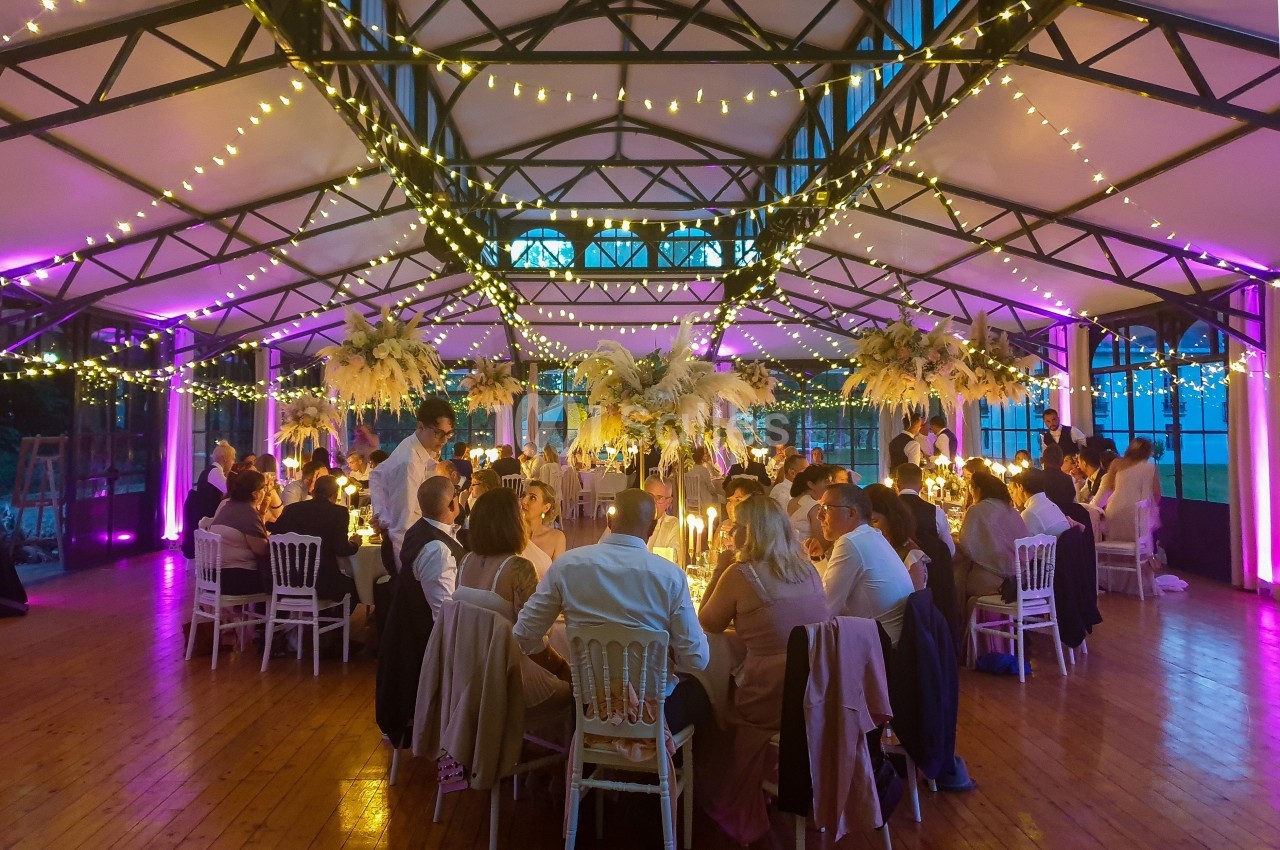 Salle de réception décorée avec des guirlandes lumineuses, des tables dressées et des invités assis lors d'un événement.