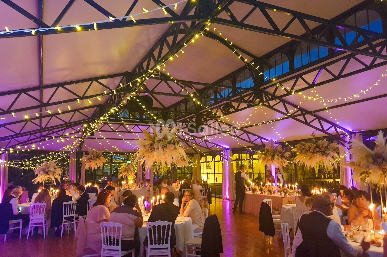 Salle de réception décorée avec des guirlandes lumineuses, des tables dressées et des invités assis lors d'un événement.