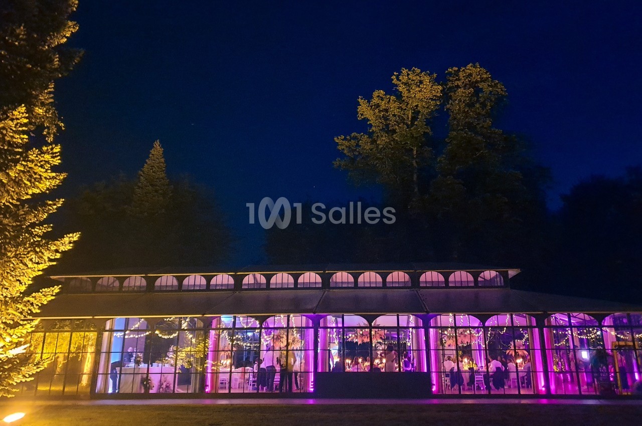 Vue nocturne d'une verrière illuminée par des lumières violettes, entourée d'arbres et d'un jardin.