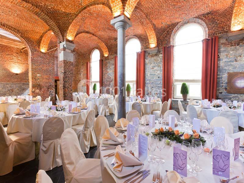 Salle de réception avec tables élégamment dressées, nappes blanches, chaises couvertes et murs en briques apparentes.