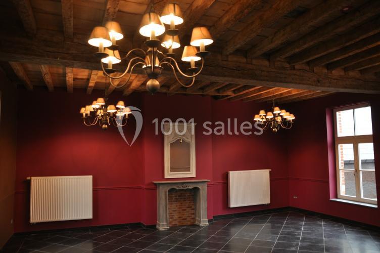 Salle avec murs rouges, plafond en poutres apparentes, lustres, cheminée décorative et fenêtres laissant entrer la lumière.