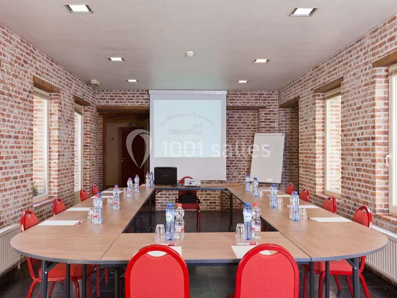 Salle de réunion avec une grande table en U, chaises rouges, bouteilles d'eau, rétroprojecteur et tableau blanc.