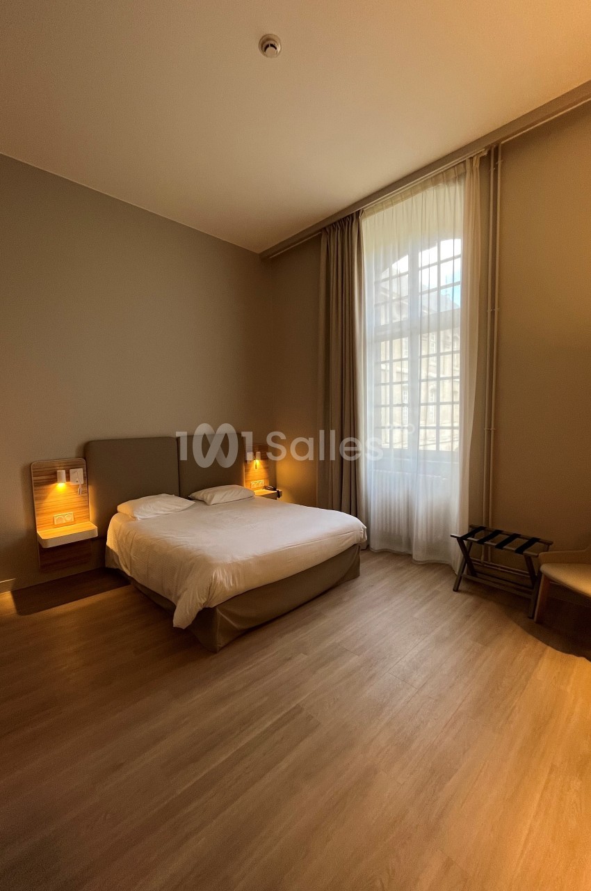 Chambre avec lit double, chevets éclairés, fauteuil, porte-bagages et grande fenêtre avec rideaux translucides.