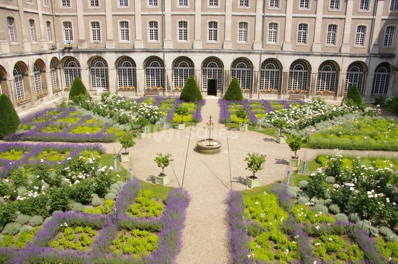 Jardin à la française avec parterres de lavande, rosiers et fontaine centrale, entouré d'un bâtiment historique.