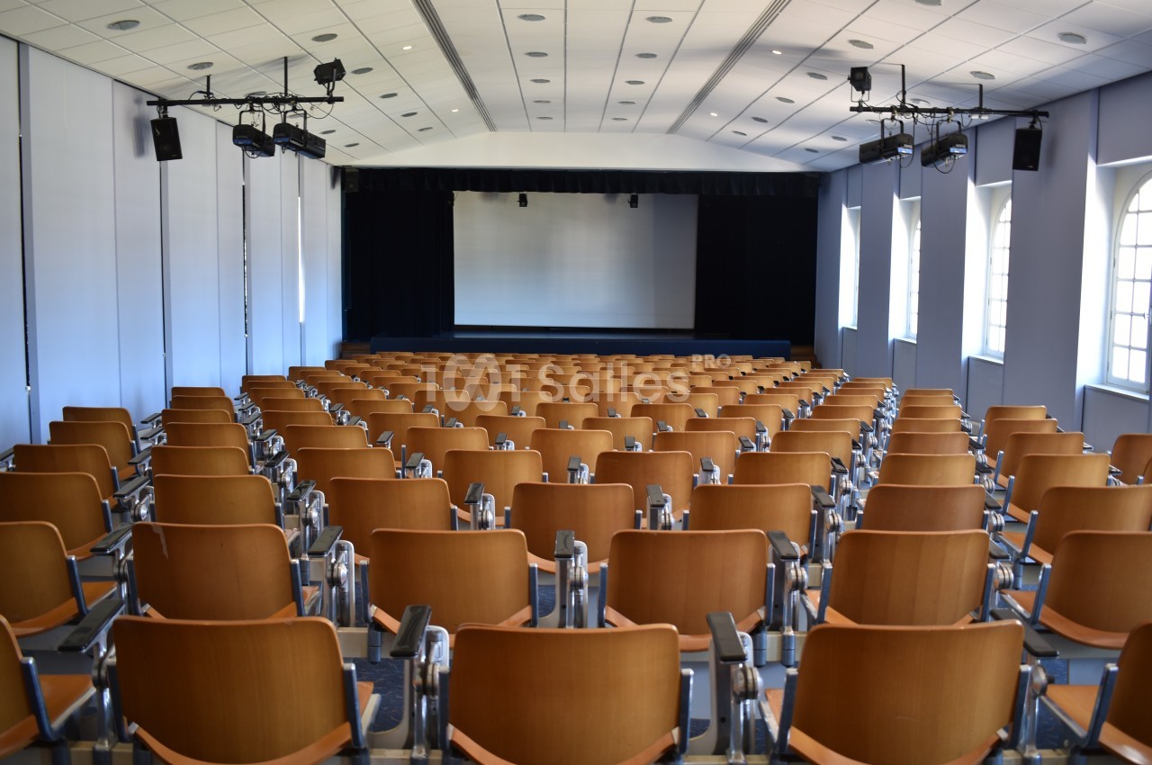 Salle de conférence vide avec des rangées de sièges en bois, un écran blanc et des projecteurs au plafond.