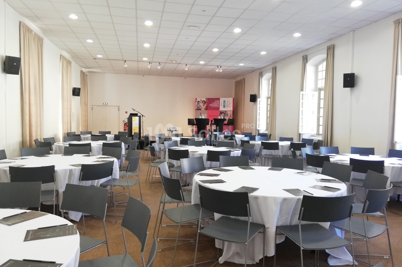 Salle de conférence lumineuse avec des tables rondes dressées de nappes blanches et des chaises grises.