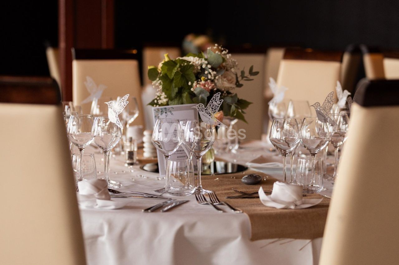 Table élégamment dressée avec nappes blanches, verres, couverts et centre de table floral dans un cadre raffiné.