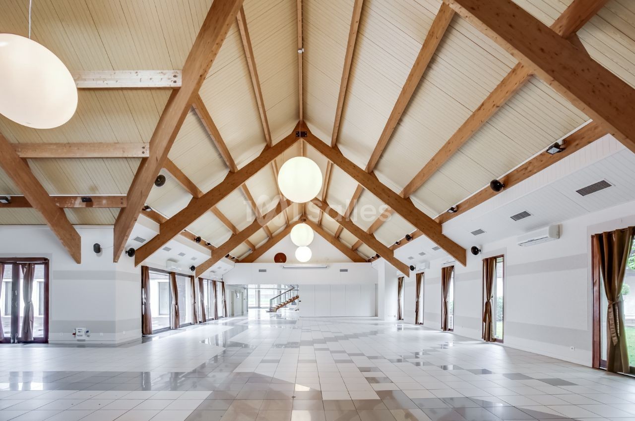 Grande salle lumineuse avec plafond en bois, poutres apparentes, carrelage clair et larges baies vitrées.
